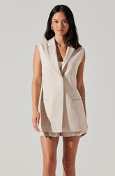A woman is wearing a sleeveless, beige, tailored vest with lapels and two front pockets. She has paired it with matching beige shorts.